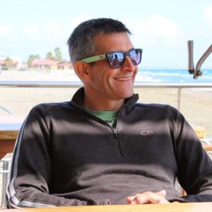 A man wearing sunglasses sitting at a table in front of the ocean.